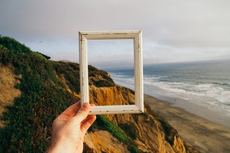 A hand holding up a photo frame before mountains and the sea